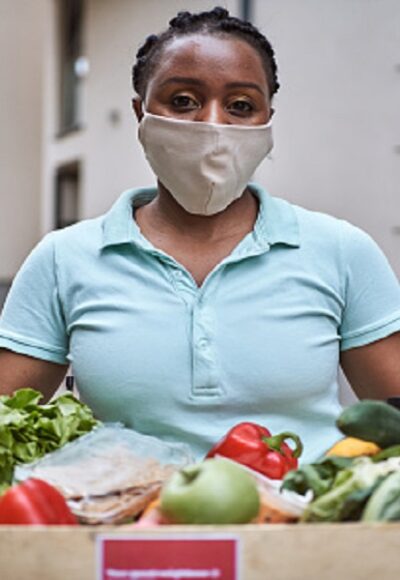 Young diligent female delivery person, Black ethnicity deliver a crate with fresh vegetable and fruits to a customer and people in need during COVID-19 pandemic while she wearing PPI