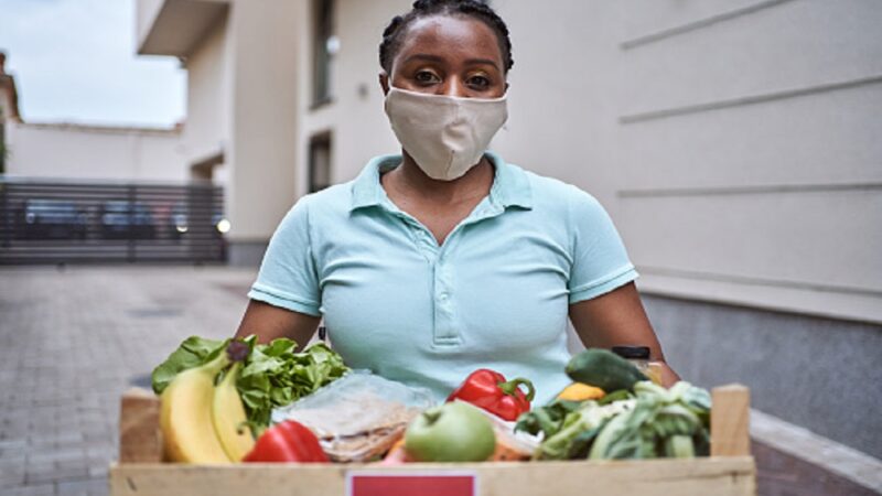 Young diligent female delivery person, Black ethnicity deliver a crate with fresh vegetable and fruits to a customer and people in need during COVID-19 pandemic while she wearing PPI