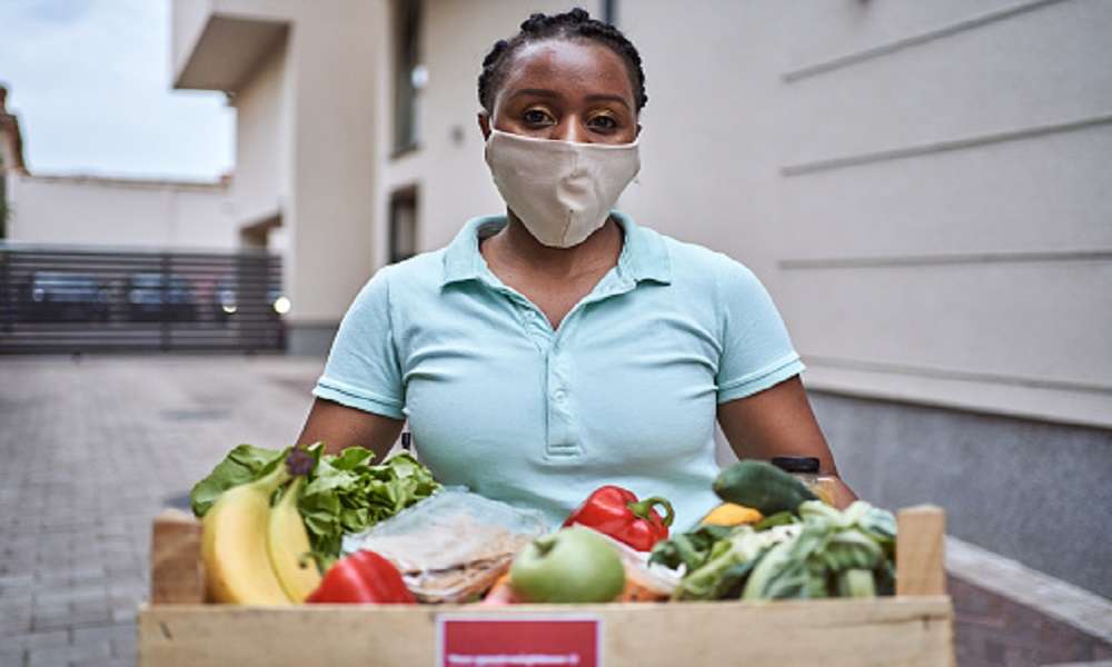 Young diligent female delivery person, Black ethnicity deliver a crate with fresh vegetable and fruits to a customer and people in need during COVID-19 pandemic while she wearing PPI